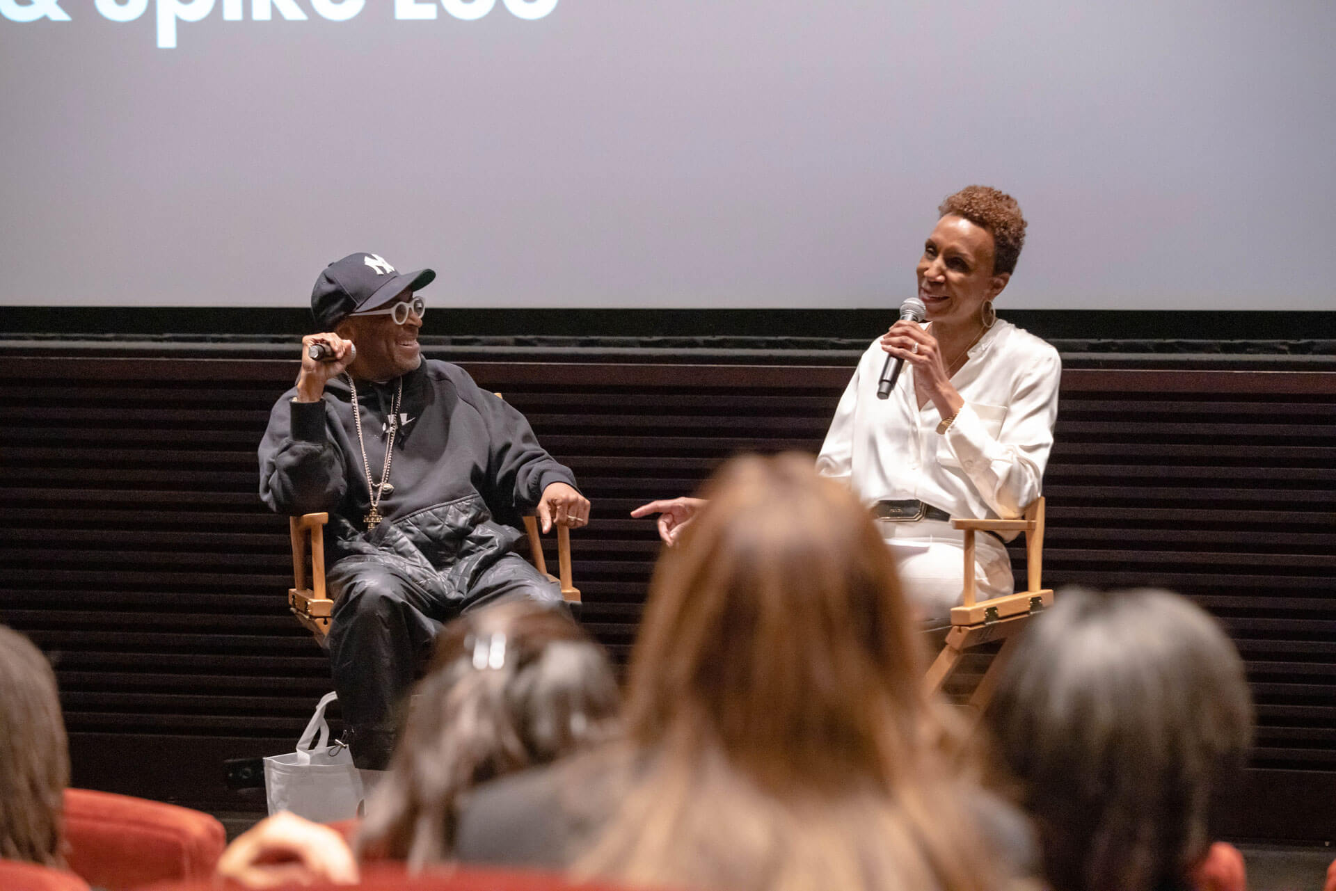A panel discussion featuring two speakers on stage, one in a casual hoodie and cap, and the other in a polished white outfit, engaging with an audience.