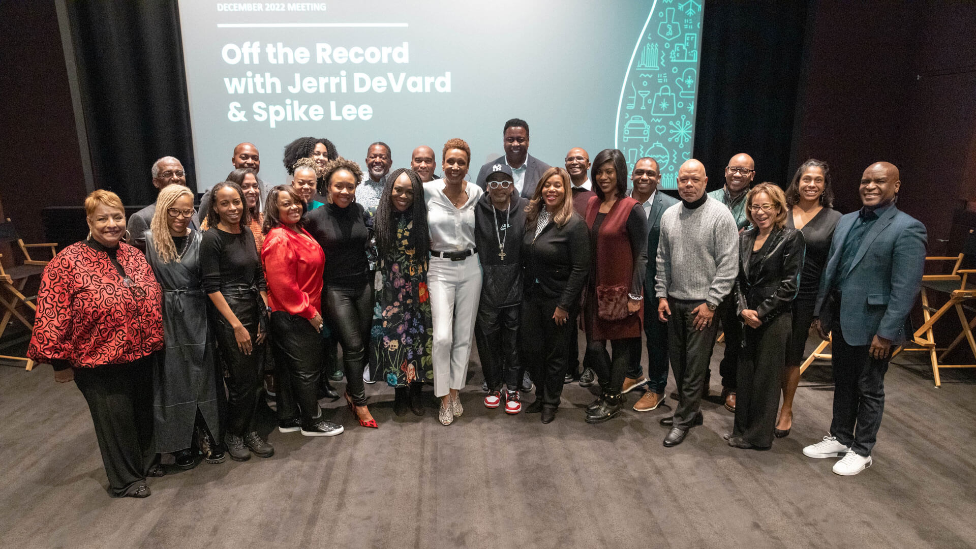 A large group photo from an event, showing a diverse group of attendees, including the speakers, standing together on a theater stage with a presentation screen in the background.