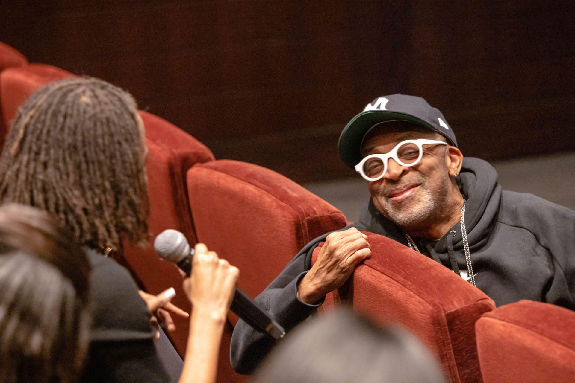 A speaker in a hoodie leans on a theater seat, smiling and interacting with another person holding a microphone during a candid moment in a discussion.