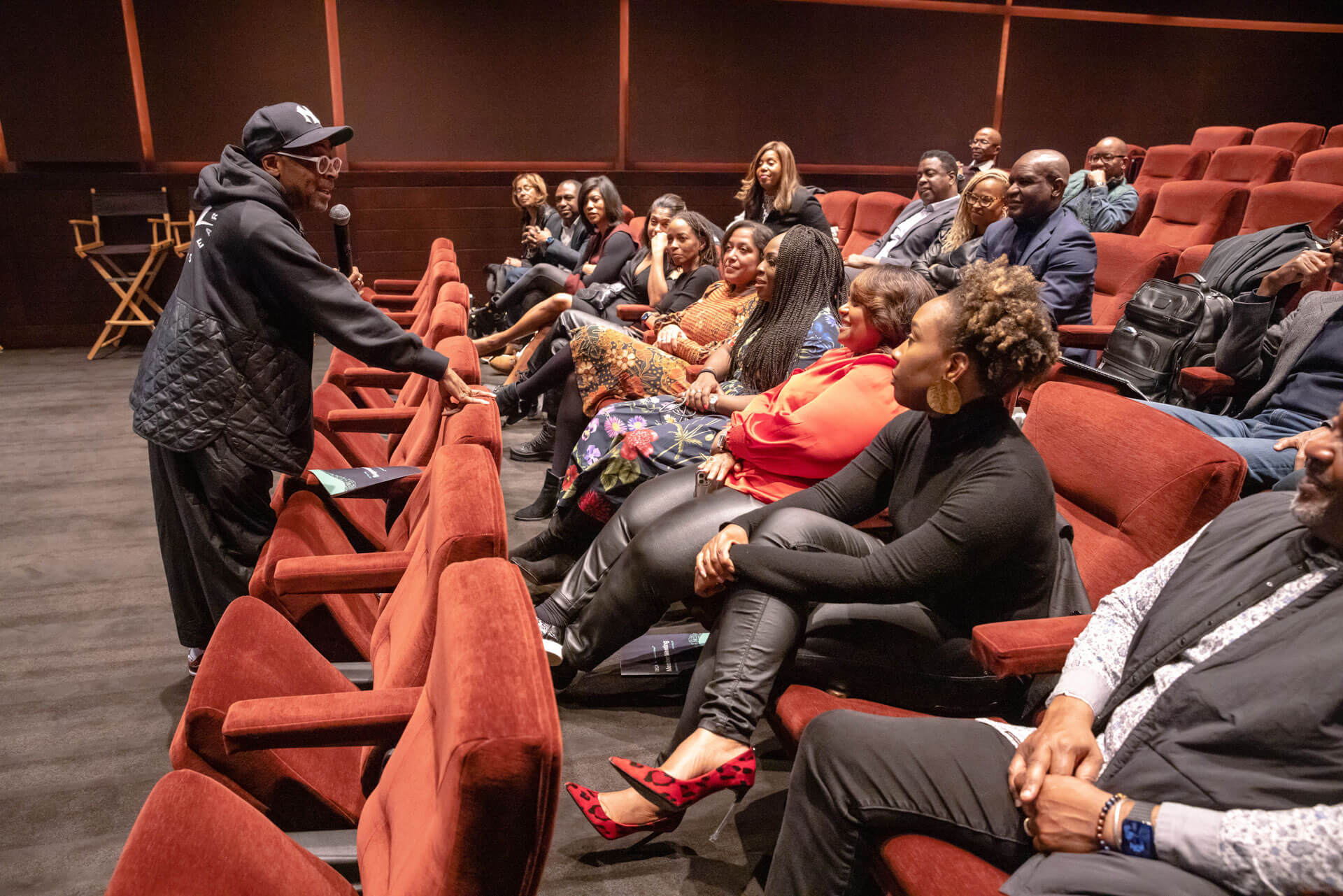 A presenter, dressed in a casual hoodie, engages a seated audience in a theater, fostering an interactive and lively discussion.