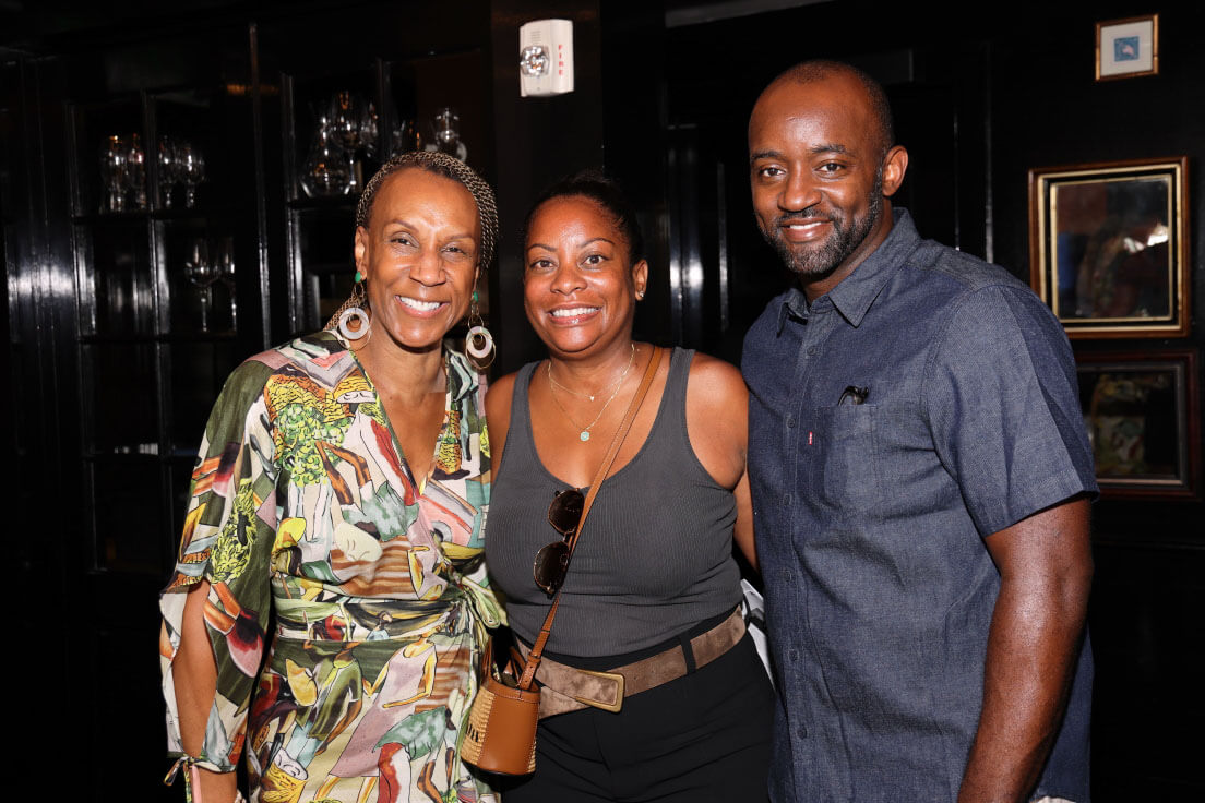Three people standing and smiling for the camera. The woman in the center wears a patterned dress, flanked by a man in a denim shirt and a woman in casual attire with a crossbody bag.