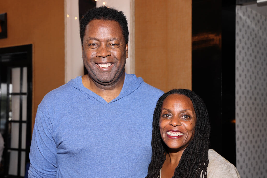 A man in a blue shirt and a woman with long dreadlocks smile together in a warmly lit indoor space.