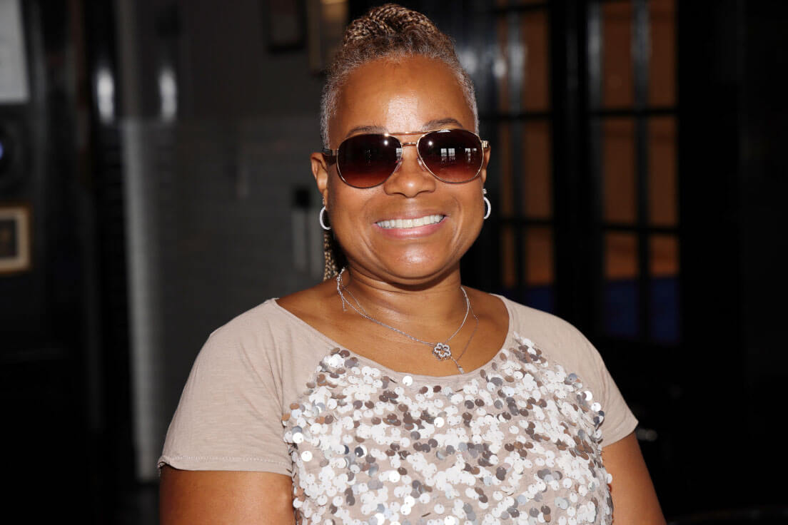 A woman wearing sunglasses and a sparkly beige top smiles warmly in an indoor setting.
