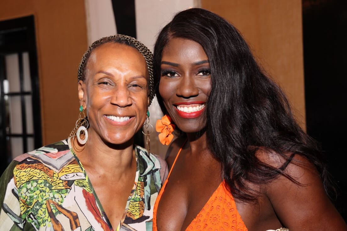 Two women smile for the camera, one in a colorful patterned dress and the other with long, flowing hair in a bright orange dress.