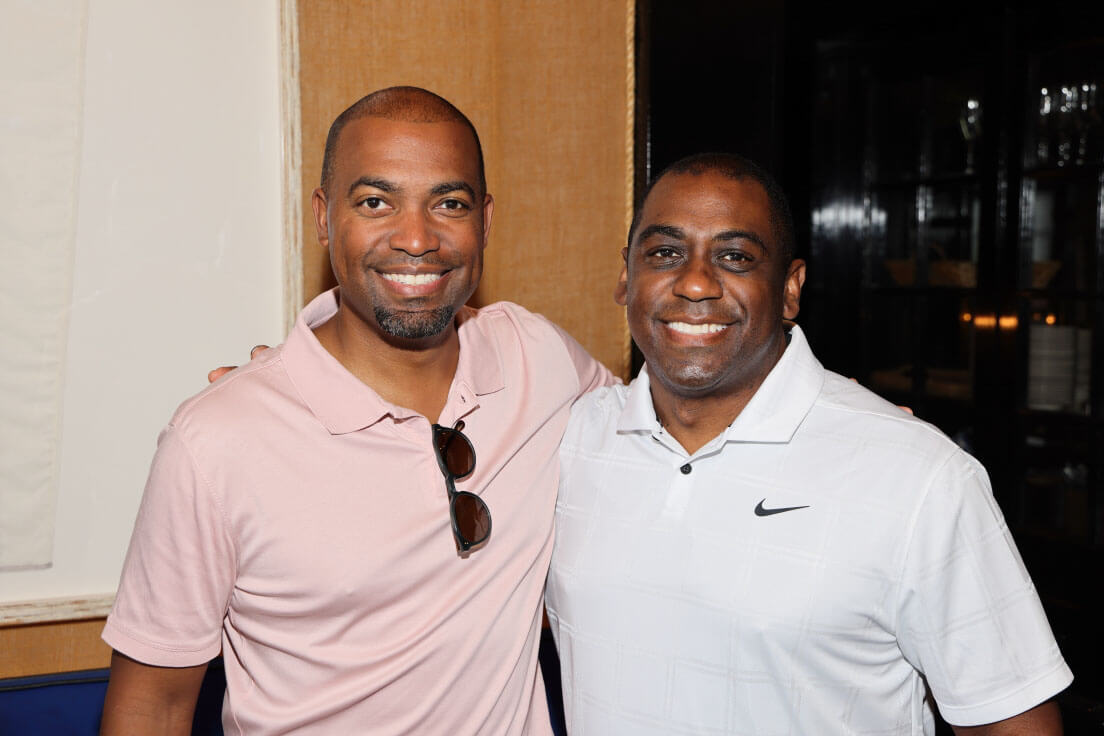 Two men standing side by side, smiling, with one in a pale pink shirt and the other in a white polo shirt with a logo.