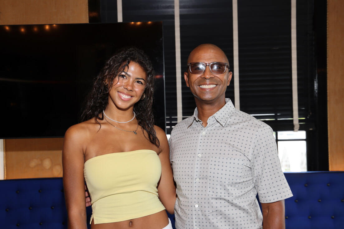 A young woman in a yellow tube top smiles beside a man in glasses wearing a patterned button-down shirt, standing in front of a dark background.