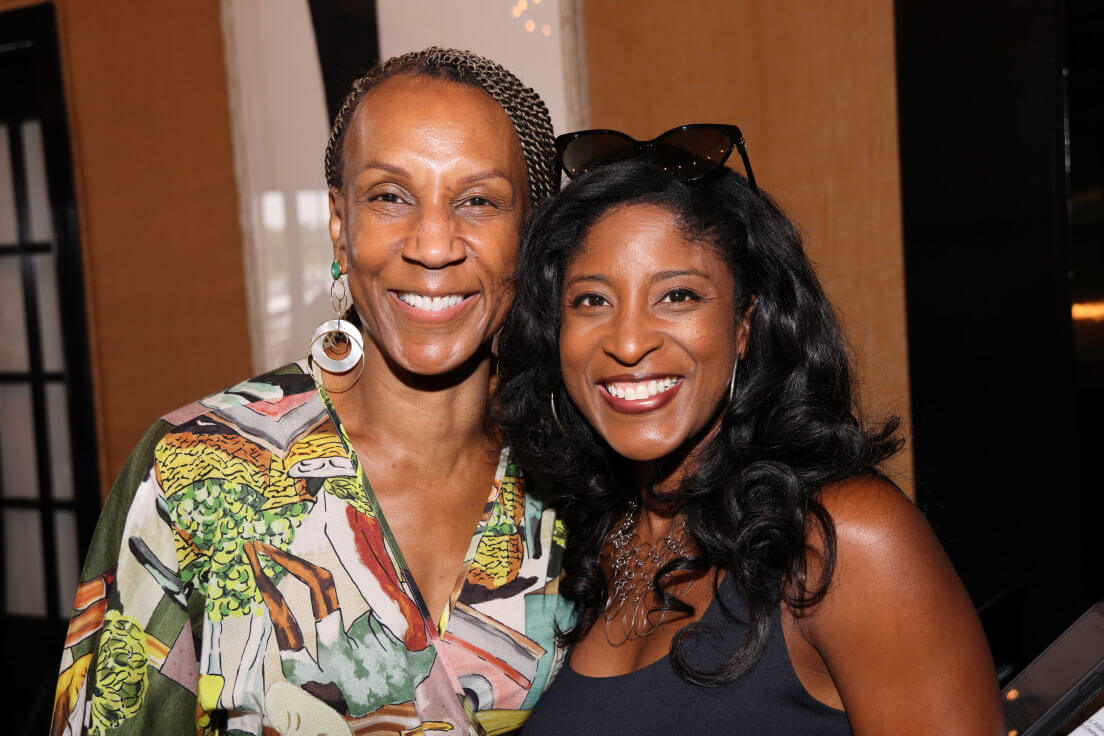 Two women smiling closely for the camera, one wearing a patterned blouse and the other with wavy hair and sunglasses on her head.