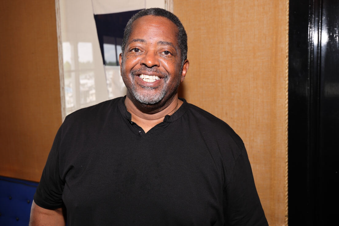A man in a black shirt smiling broadly, standing in a warmly lit indoor space with framed decorations on the wall behind him.