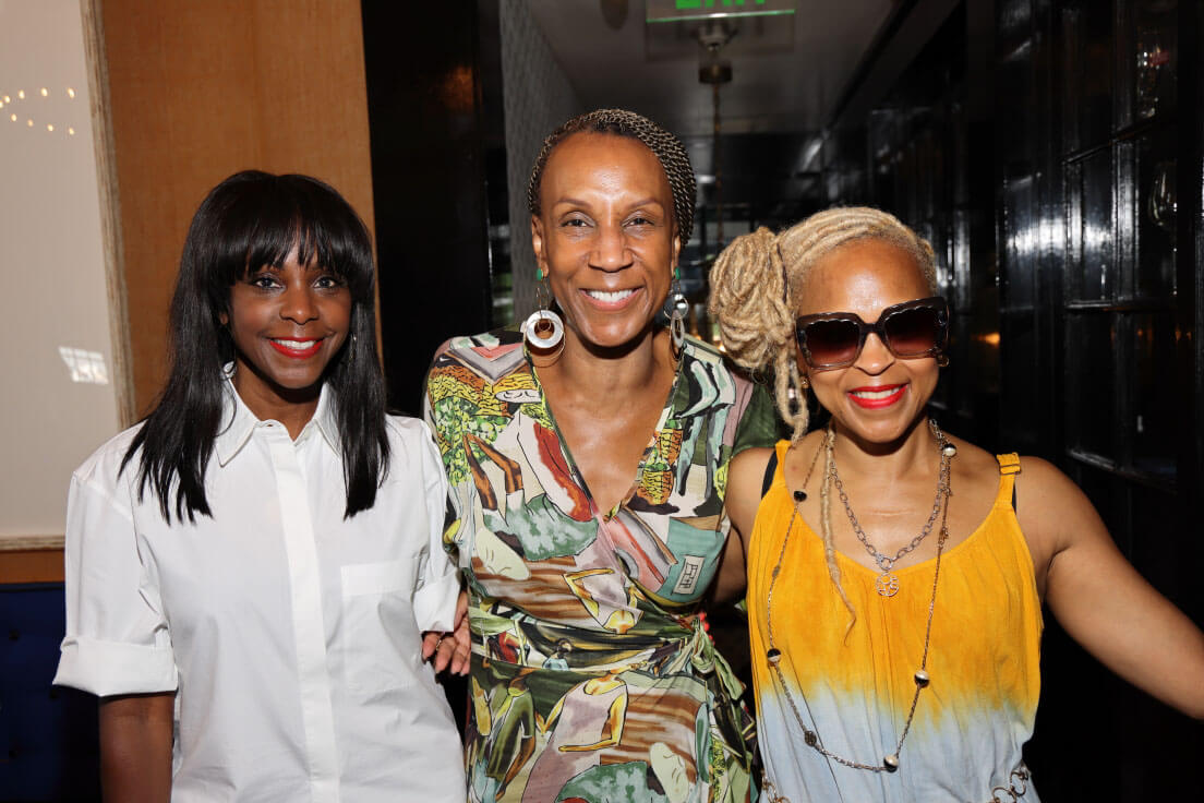 Three women posing together with cheerful expressions. One wears a white shirt, another a colorful patterned top, and the third a yellow dress with sunglasses.