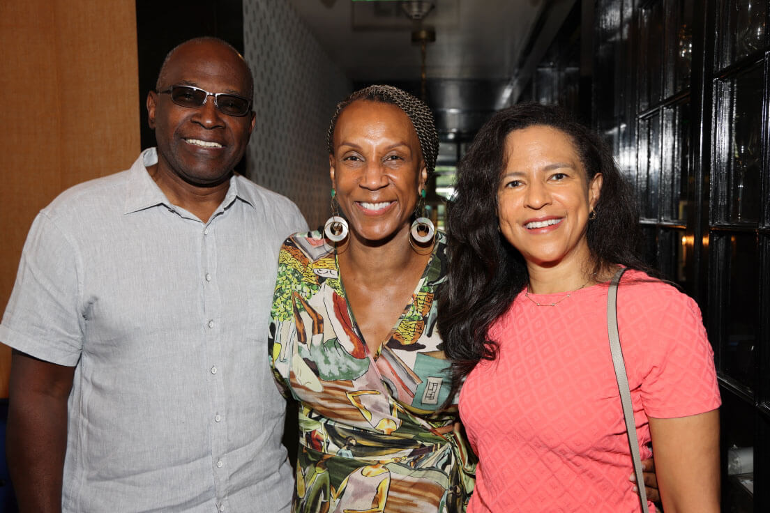 Two women and a man standing together and smiling warmly. The man is in a light grey shirt, one woman in a patterned blouse, and the other in a pink top.