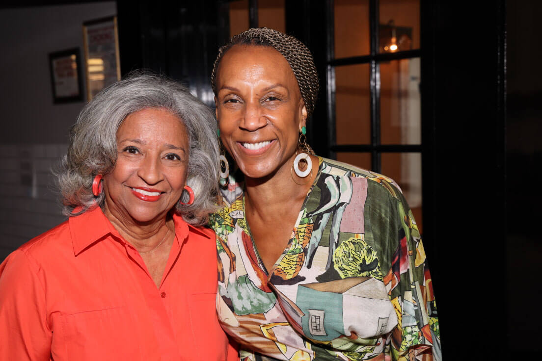 Two women, one with grey hair in an orange blouse and another with braids in a colorful patterned top, smiling side by side in a well-lit space.