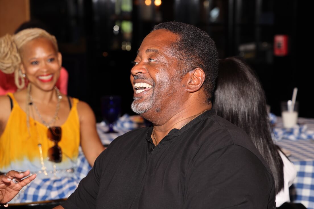 A man in a black shirt laughs heartily while a woman in a yellow top smiles in the background, seated at a table with checkered cloth.