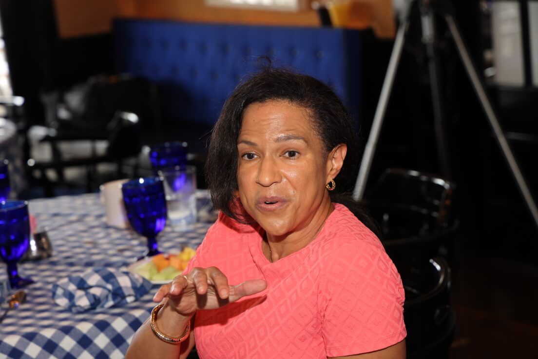 A woman in a bright pink top gestures expressively while seated at a table set with checkered cloth and blue glasses.