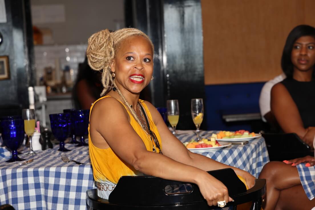 A woman with blonde locs smiles warmly while seated at a table with a checkered tablecloth, surrounded by glasses and plates of fruit in a cozy indoor setting.
