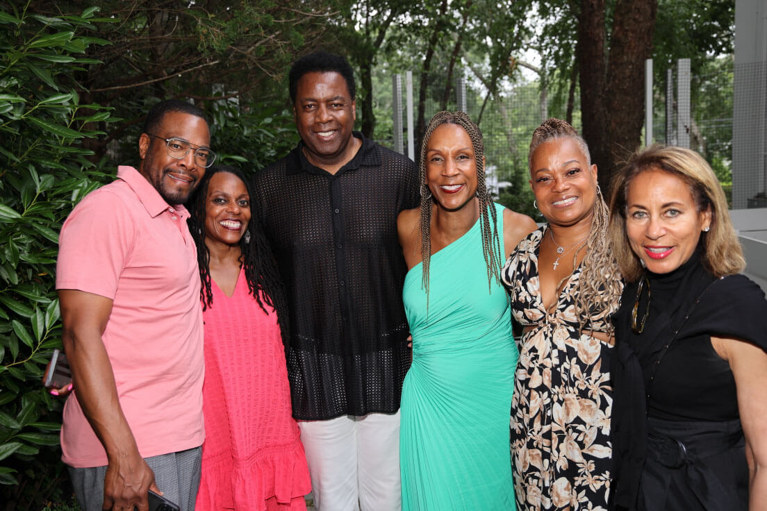 A group of six people smiling and posing together outdoors, wearing a mix of casual and formal attire, with greenery and a modern building in the background.