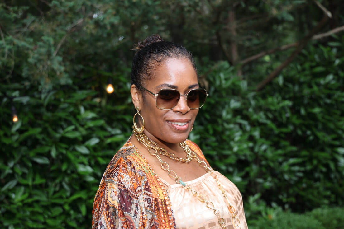 A woman with short hair and sunglasses smiling, wearing a colorful patterned outfit with bold jewelry, posing in front of a green, wooded backdrop.