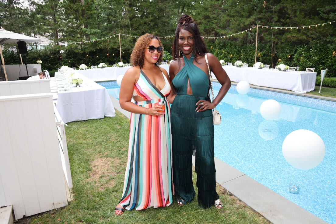 Two women posing together by a pool, one in a colorful striped dress and the other in a teal halter jumpsuit, with decorated tables and trees in the background.
