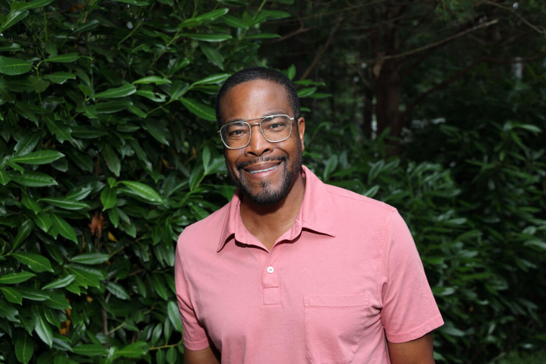 A man in a light pink shirt and glasses smiling outdoors, with greenery softly blurred in the background.
