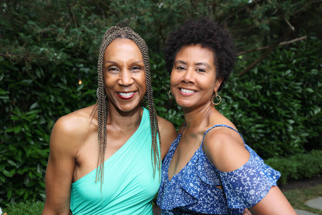 Two women smiling together outdoors, one in a green one-shoulder dress and the other in a blue floral off-shoulder dress, with greenery in the background.