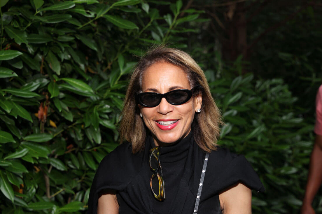 A woman with shoulder-length blonde hair, wearing black sunglasses and a black outfit, smiling outdoors with greenery in the background.