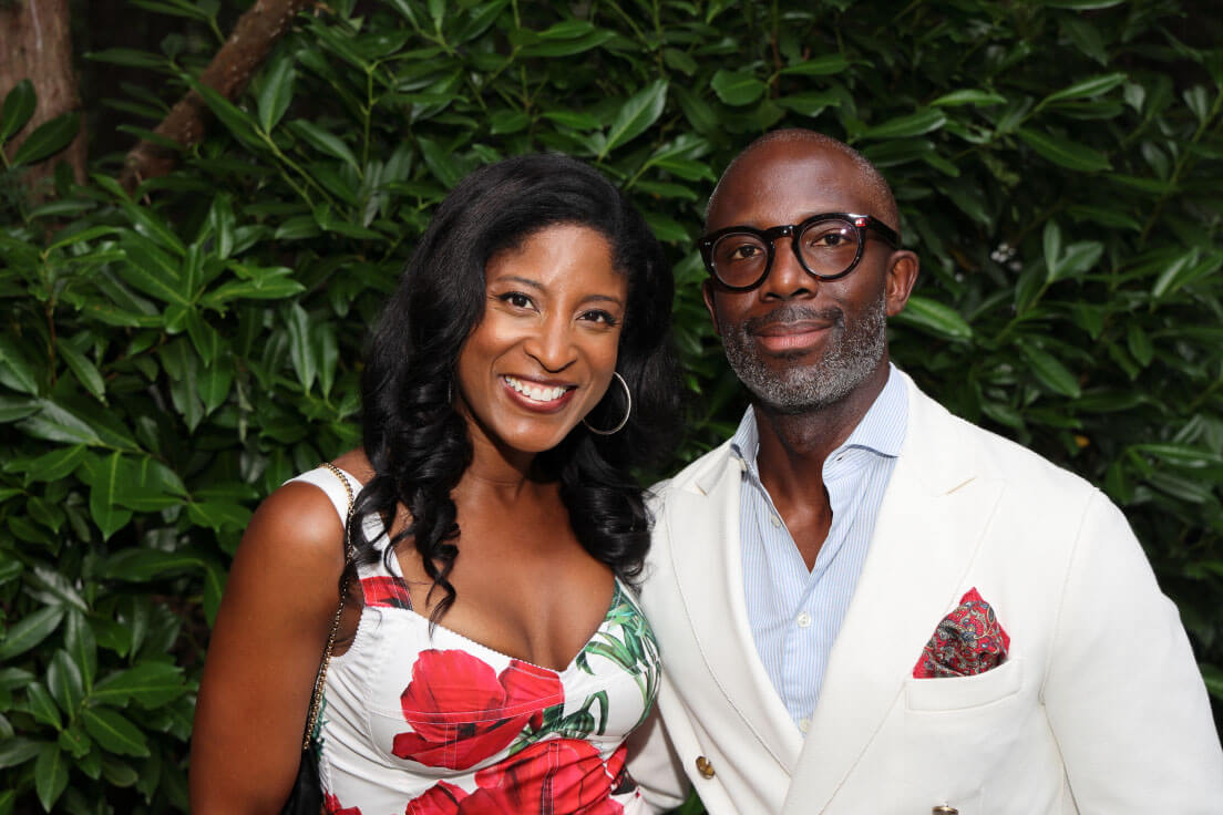 A man and a woman posing together; the woman wears a floral dress, and the man is in a white blazer with glasses and a pocket square, standing in front of greenery.