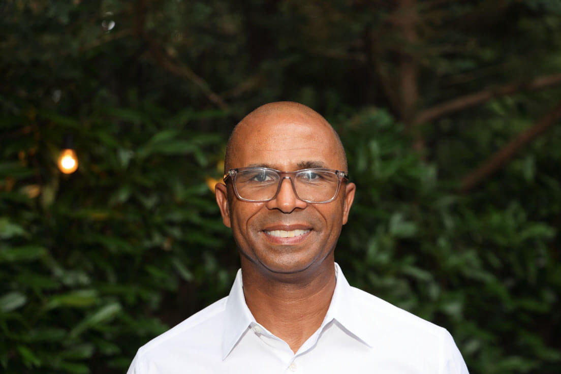 A close-up of a man wearing glasses and a white shirt, smiling with soft greenery in the background.