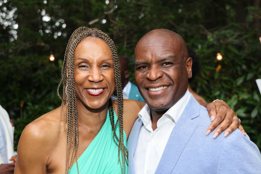 A man and a woman posing together, smiling widely; the man wears a light blue blazer, and the woman wears a green, one-shoulder dress, with greenery in the background.