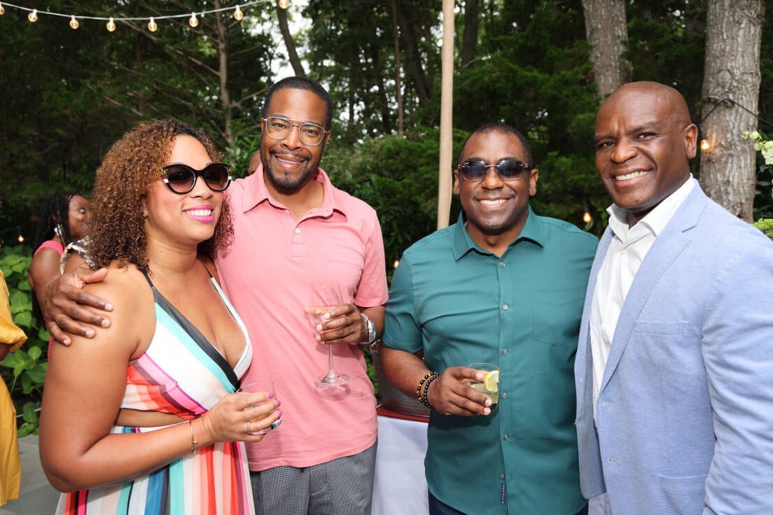 A group of four individuals posing outdoors, two women and two men; one woman in a colorful striped dress and sunglasses, and the men in casual shirts.