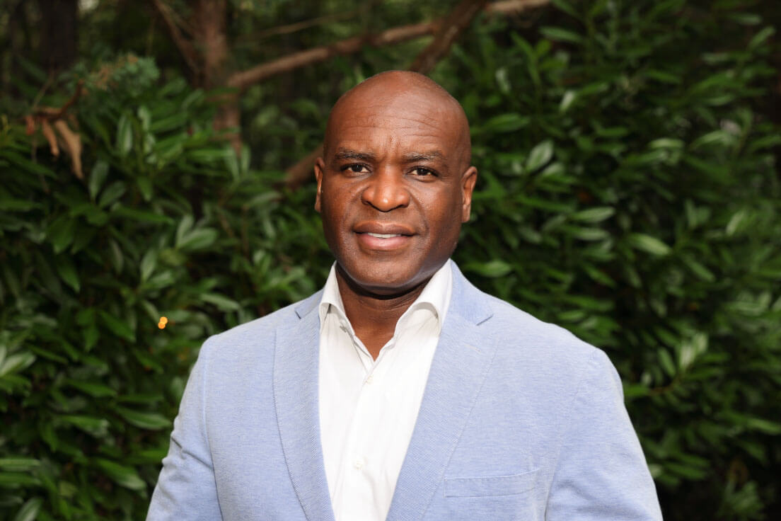 A man in a light blue blazer and white shirt smiling, standing outdoors with greenery in the background.