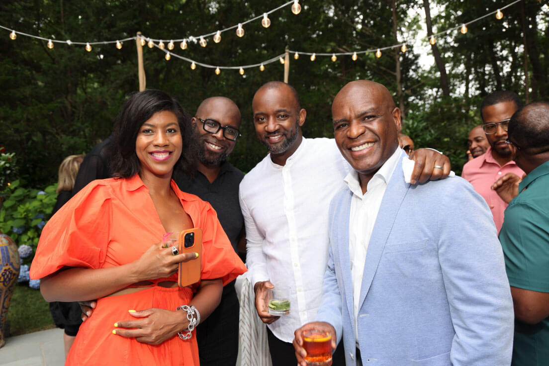 A group of four individuals posing and smiling, enjoying a casual outdoor event with string lights above.