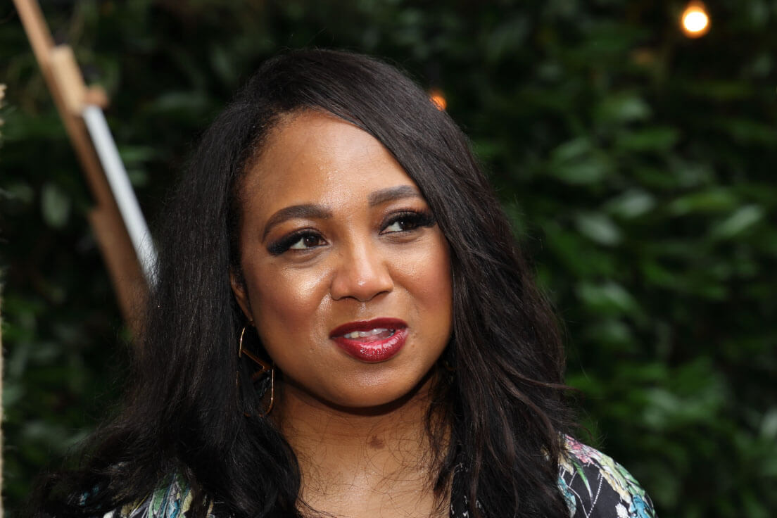 A close-up of a woman with long dark hair, wearing a floral outfit, with greenery softly blurred in the background.