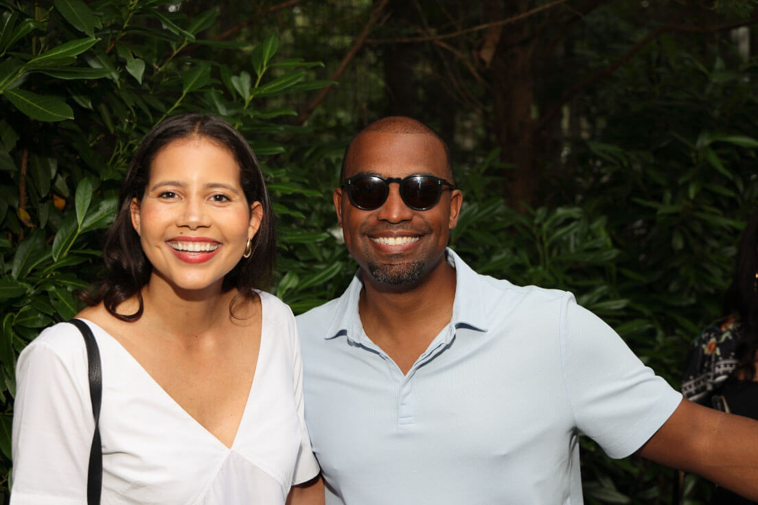 A man and a woman smiling together, the man wearing sunglasses and a light blue polo shirt, and the woman in a white blouse.