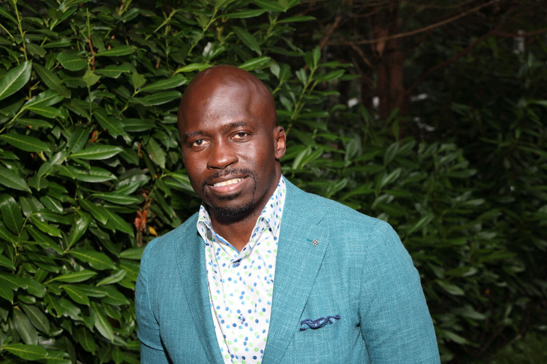 A man in a teal blazer with a patterned shirt smiling, standing outdoors with lush greenery as a backdrop.