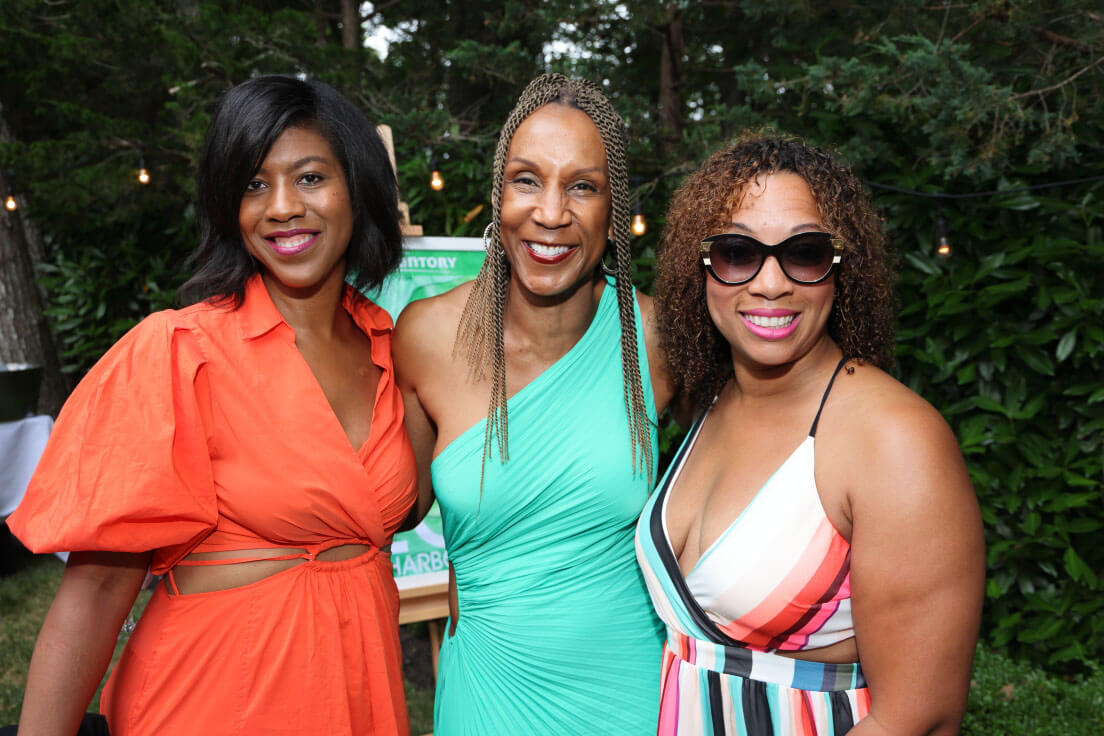 Three women posing together, each in bright, colorful attire, at an outdoor gathering with trees in the background.