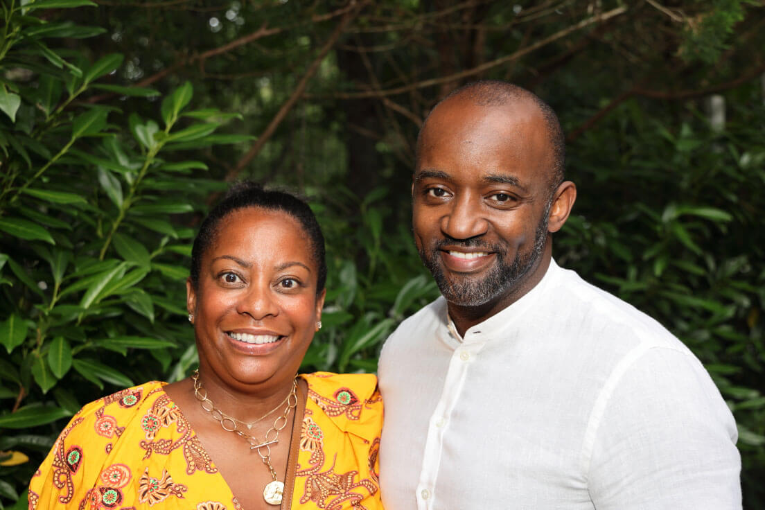 A man and a woman smiling side by side, with the woman wearing a vibrant yellow outfit and the man in a white shirt.