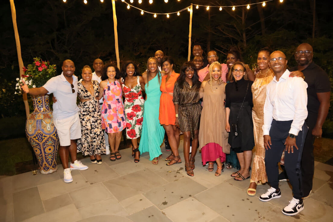A large group of people posing together outdoors at night, smiling under string lights. The atmosphere is festive, with attendees dressed elegantly.