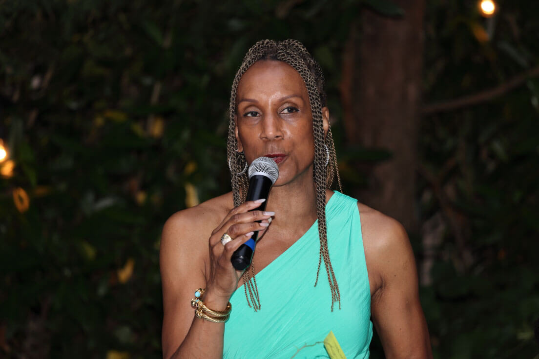 A woman in a green one-shoulder dress speaks into a microphone during an outdoor evening event. The background features greenery and soft lighting.