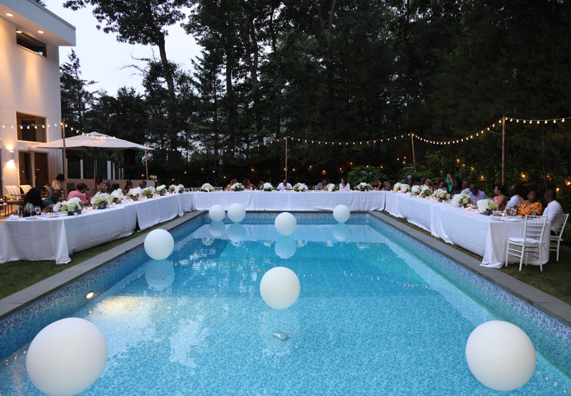 A beautifully set poolside dinner table at twilight, with white tablecloths, floral centerpieces, and glowing orbs floating in the pool.