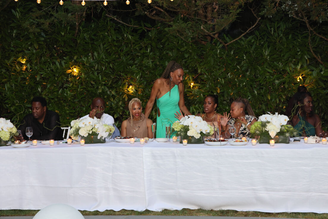 Guests seated at a long, elegantly decorated outdoor table under string lights, enjoying a meal and conversation in a lush garden setting.
