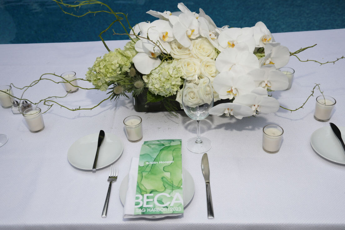 A close-up of a beautifully arranged place setting at an outdoor dinner, featuring white orchids, candles, and custom event menus.