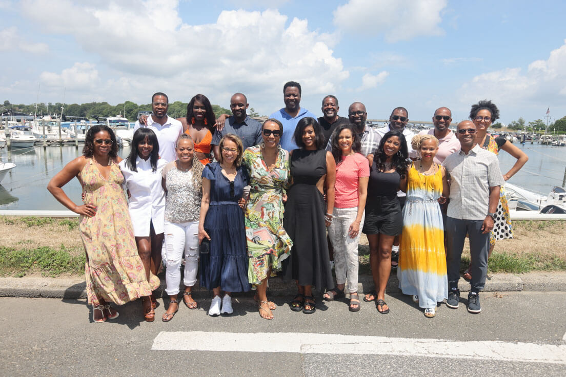A large group of individuals posing together in a sunny outdoor setting by a marina. Everyone is smiling, dressed in a mix of casual and semi-formal attire.
