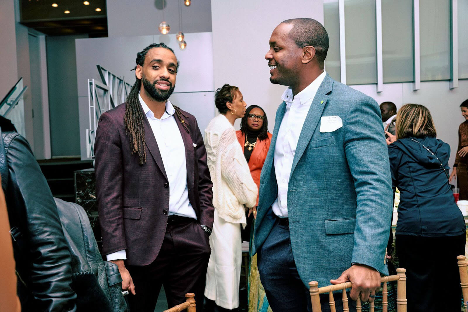Two men chatting casually, smiling warmly at each other, as others mingle in the background at a networking event.