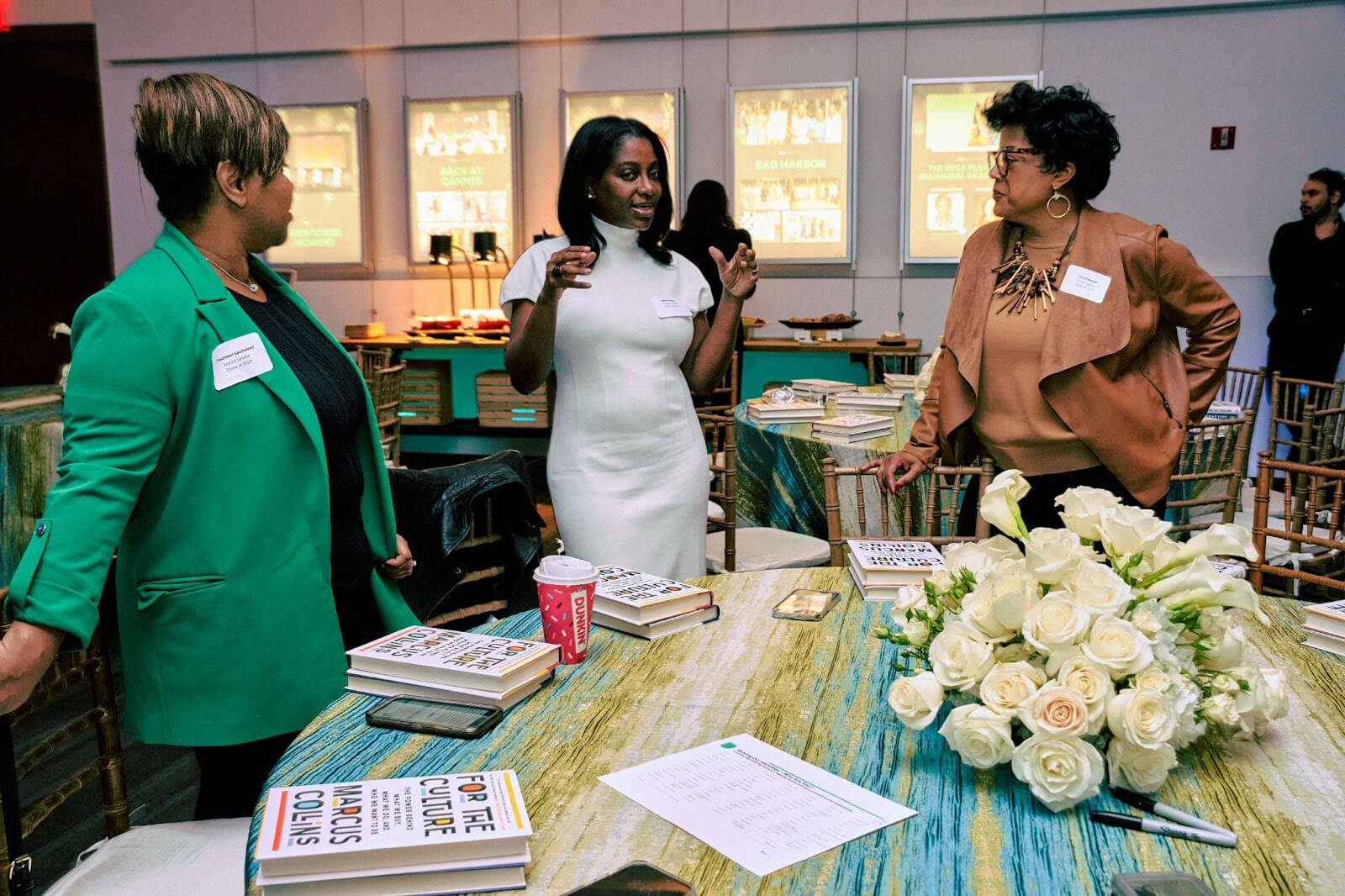 Three women standing at a table, engaged in an animated discussion, with books and flowers prominently displayed.