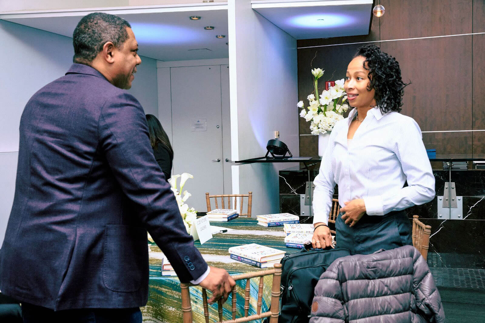 A man and a woman having a conversation near a table, with event materials and flowers in the background.