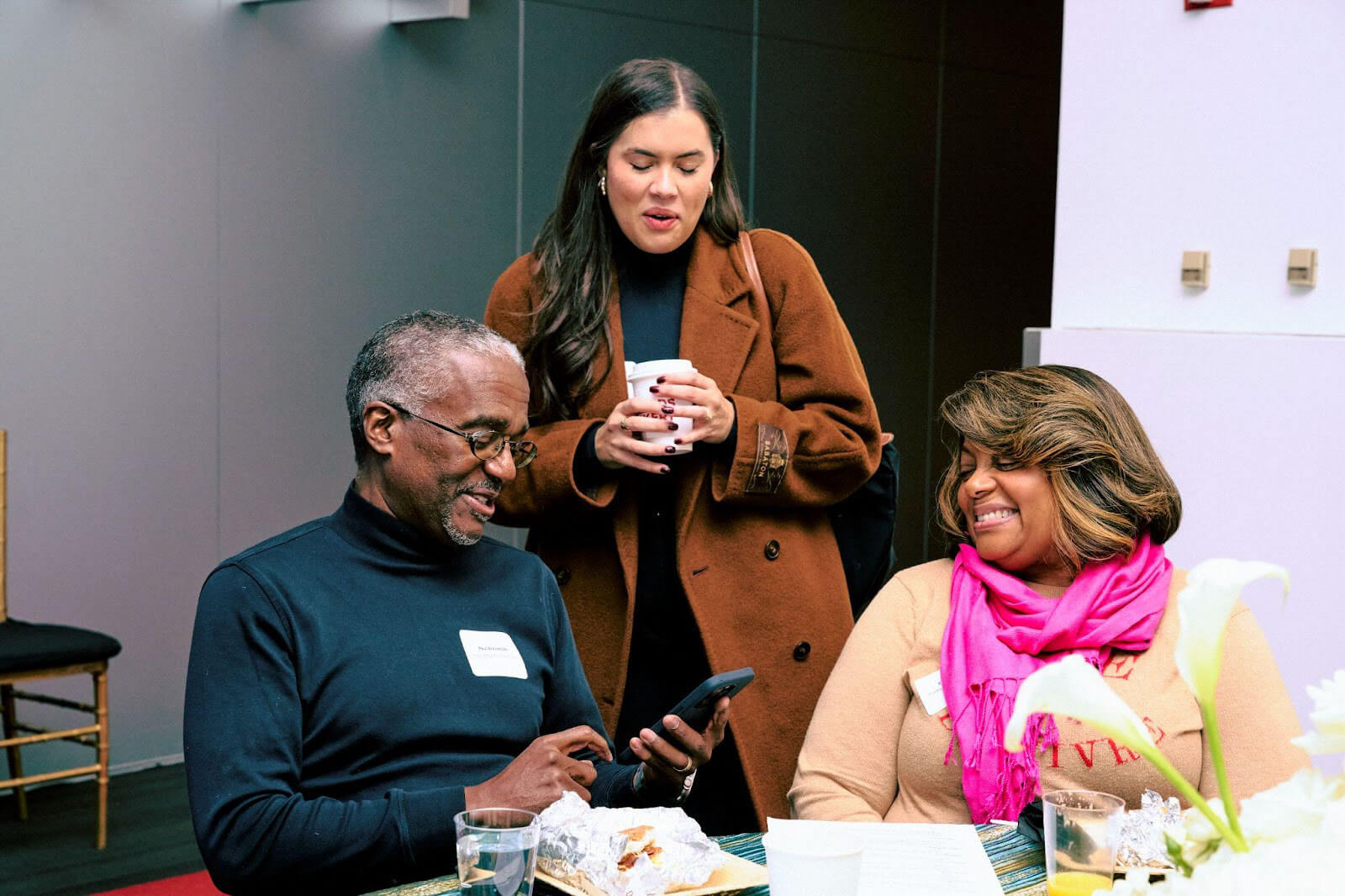 A lighthearted moment where a man shows something on his phone, while two women stand nearby, one laughing and the other holding a cup.