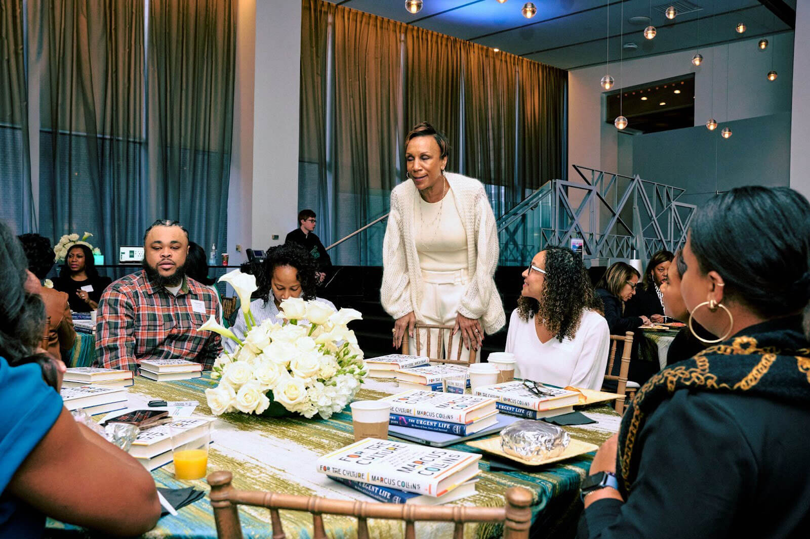 A woman standing and speaking to attendees at a table adorned with flowers and books during an event.