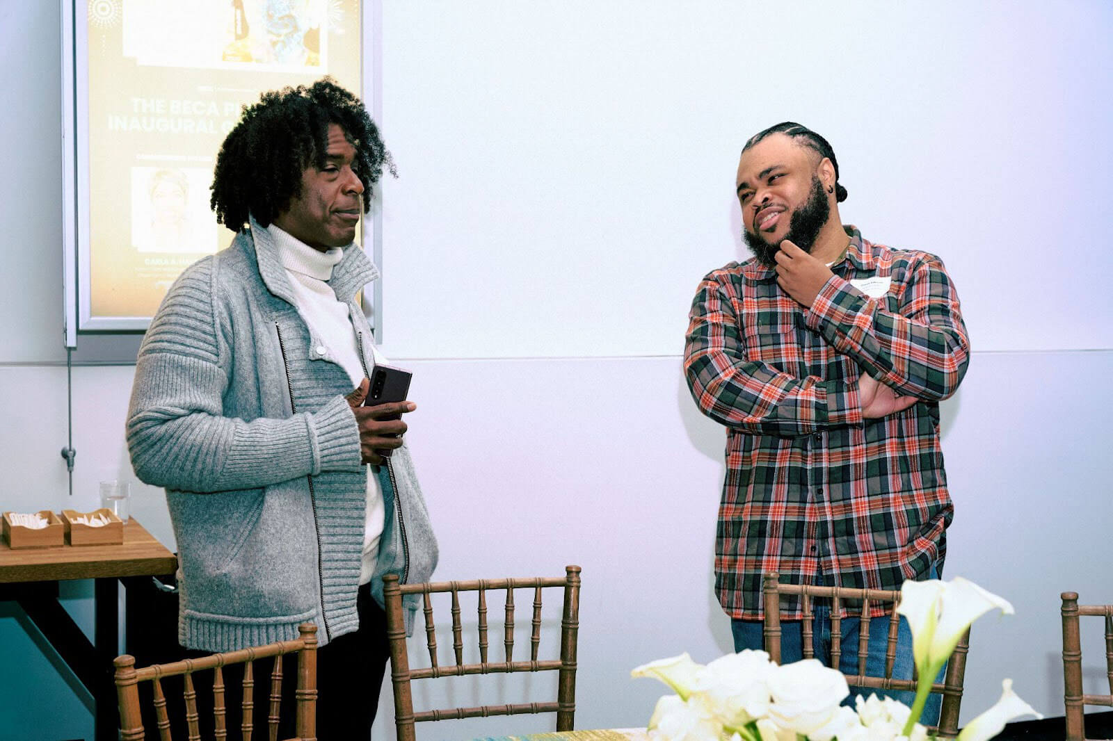 Two men in a casual conversation near a table, one holding a phone while the other listens thoughtfully.