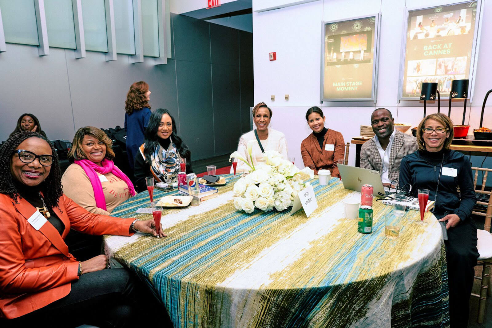 A group of seven individuals, including men and women, seated around a decorated round table with drinks, flowers, and refreshments, smiling warmly for the camera at a professional event.