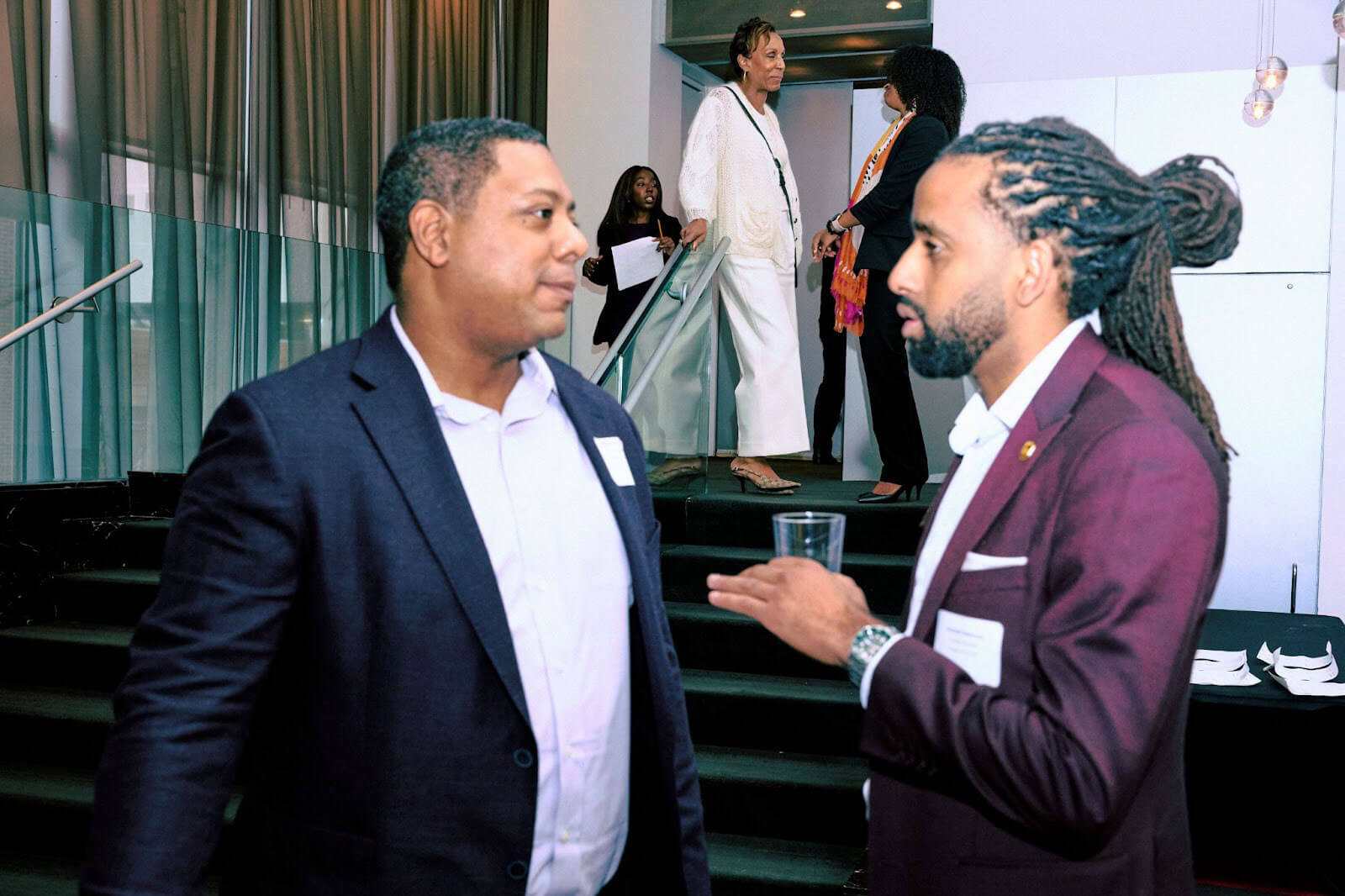 Two men in formal attire having a focused conversation near a staircase at an event, while other attendees can be seen interacting in the background.