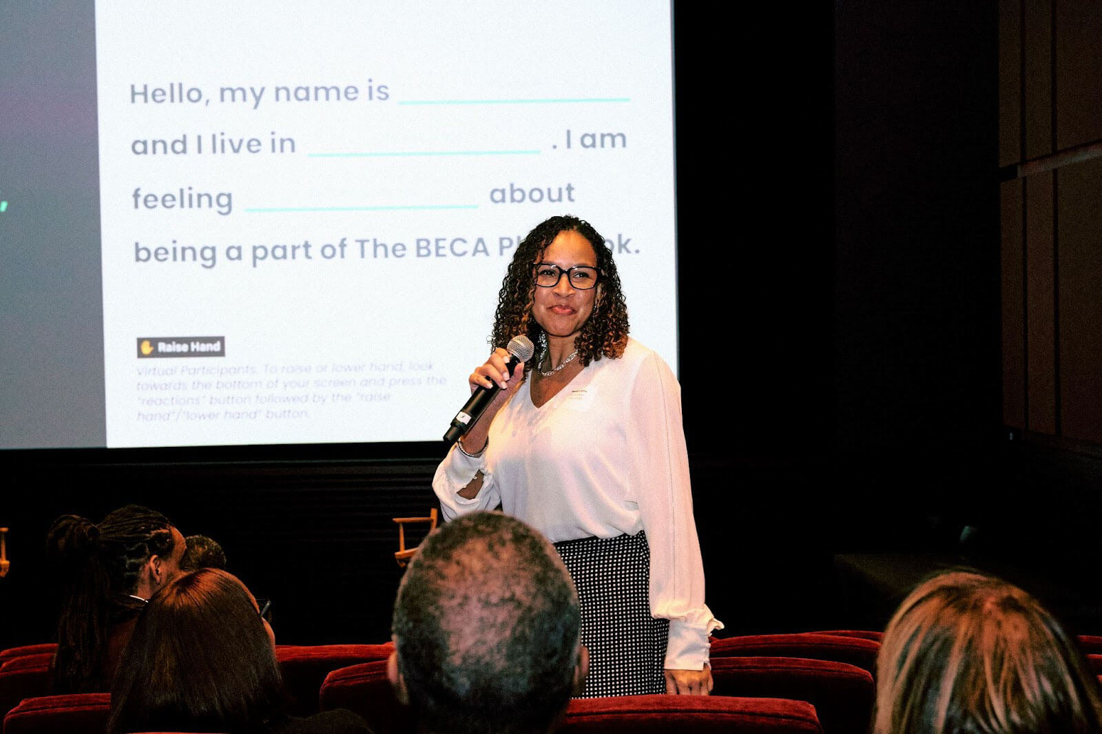 A woman standing in front of an audience with a microphone in hand, smiling and engaging with the attendees as a presentation slide is projected in the background.